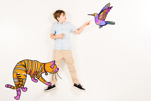 Draufsicht eines überraschten Kindes, das mit dem Finger auf einen Kolibri in der Nähe einer orangefarbenen Katze auf Weiß zeigt  - Foto, Bild