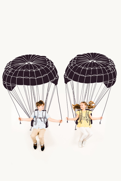 Draufsicht von fröhlichen Jungen und glücklichen Mädchen, die Fallschirme halten, während sie auf Weiß fliegen  - Foto, Bild