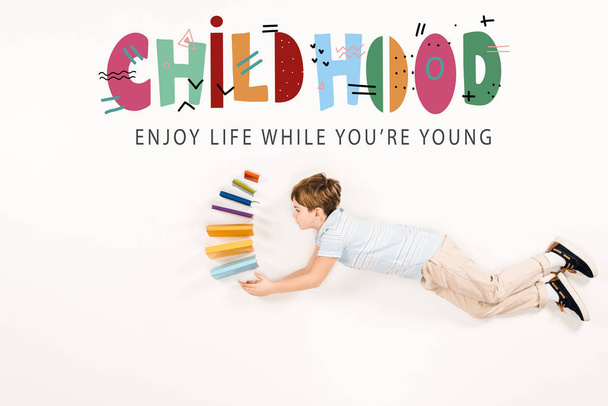 top view of kid holding books and flying near childhood enjoy life while you're young lettering on white - Photo, image