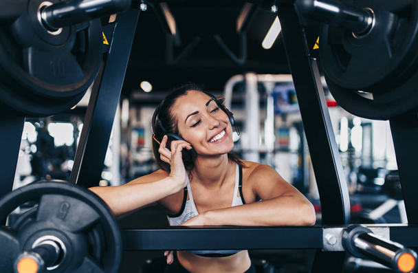 Young fit and attractive woman working out in modern gym and listening to music with bluetooth headphones and smart phone. - Photo, image