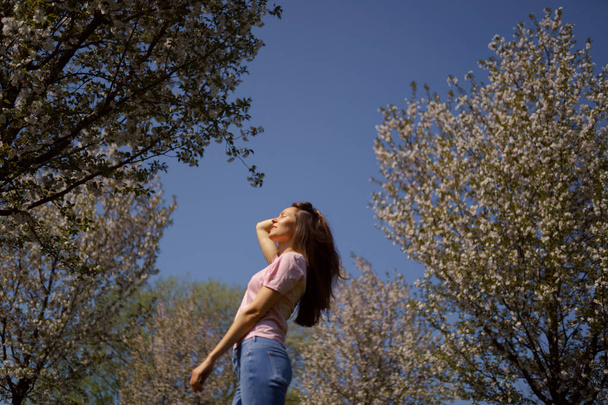 Successful business woman enjoys her leisure free time in a park with blossoming sakura cherry trees wearing jeans, pink t-shirt and long hair from behind back - 写真・画像