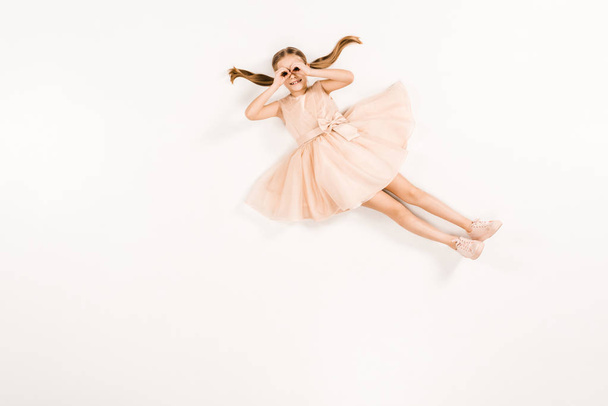 cheerful kid in dress with hands near eyes while lying on white  - Foto, Bild