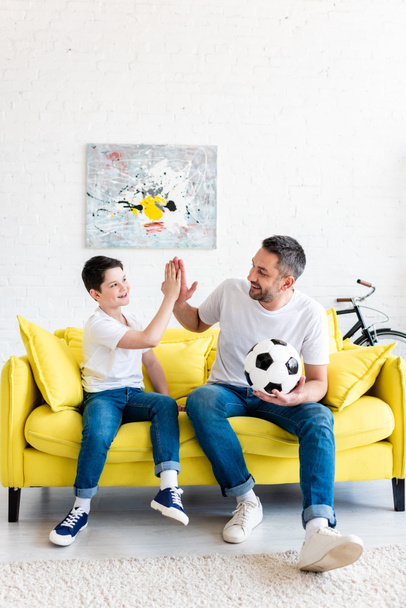 Ojciec i syn robi wysokie pięć znak siedząc na kanapie z piłką nożną w domu - Zdjęcie, obraz