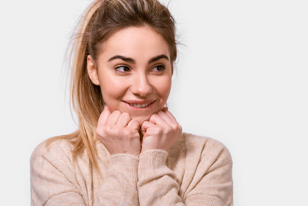 Close up portrait of cute kind young woman smiling and keeps hands on chin, looking aside, dressed beige sweater, isolated over white background. People, emotions, feeling and body language concept - 写真・画像