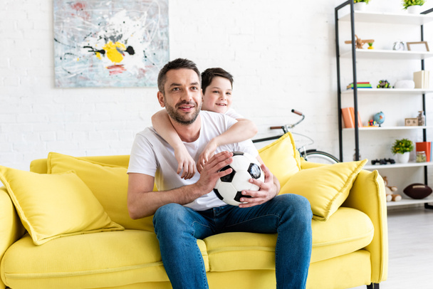 father and son watching sports match on couch with soccer ball at home - Foto, Bild