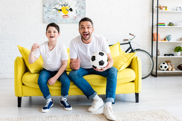 father and son cheering while watching sports match on couch with soccer ball at home - Foto, immagini