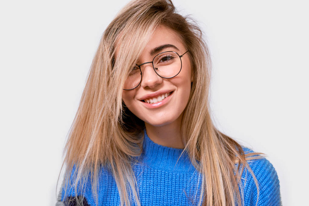 Pretty positive European young woman in stylish loose sweater and round transparent eyewear, smiling broadly and looking to the camera, isolated over white background. People emotion - Valokuva, kuva