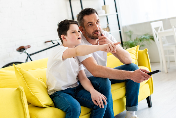 Sohn zeigt mit dem Finger, während er auf Couch sitzt und mit Vater zu Hause fernsieht - Foto, Bild