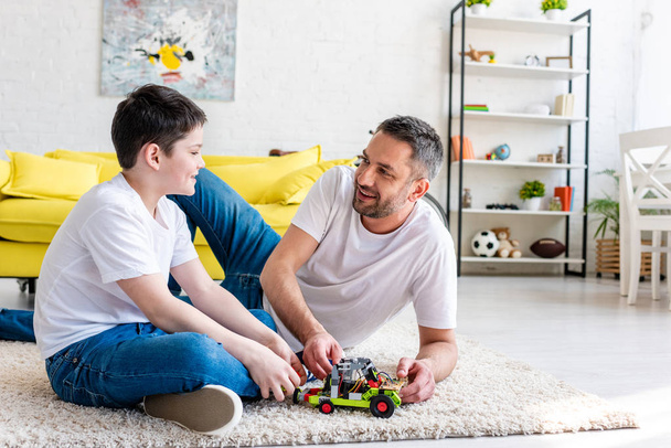 smiling father and son sitting on carpet and playing with toy car at home - Φωτογραφία, εικόνα