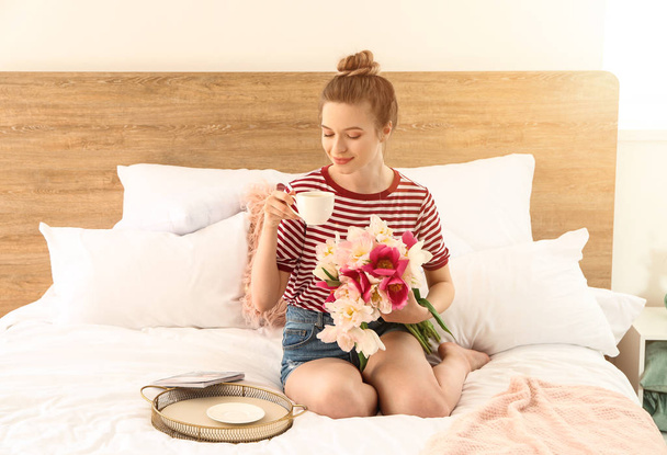 Young woman with bouquet of beautiful tulips and cup of coffee sitting on bed - Foto, Bild