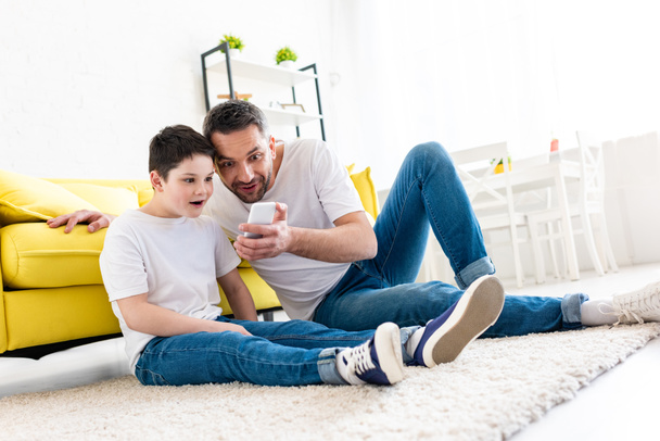 surprised father and son sitting on carpet and using smartphone in Living Room - 写真・画像