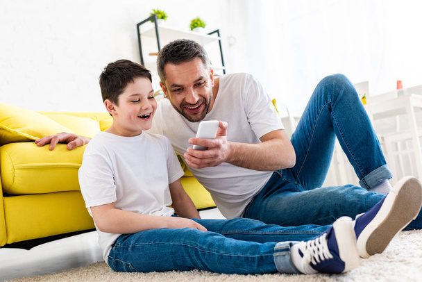 onnellinen isä ja poika käyttävät älypuhelinta olohuoneessa
 - Valokuva, kuva