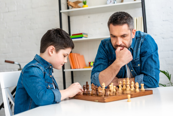 pensive father and son in denim playing chess while sitting at table at home - Fotoğraf, Görsel