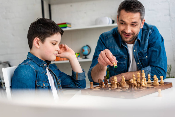 selective focus of father and son in denim playing chess while sitting at table at home - 写真・画像