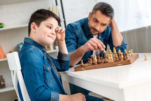 father and son in denim playing chess while sitting at table at home - Photo, Image