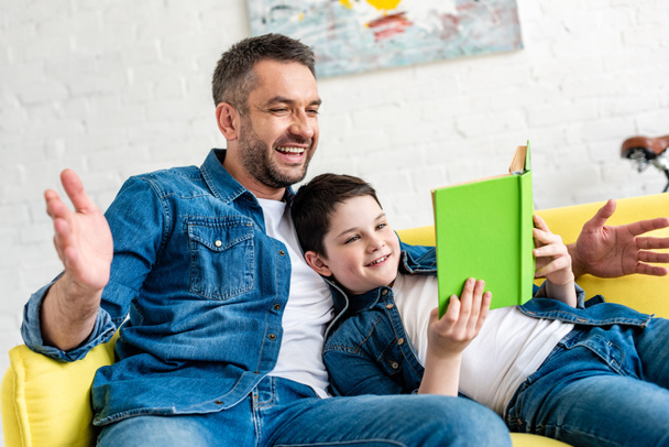 smiling father and son in denim reading book on couch at home - Foto, Bild