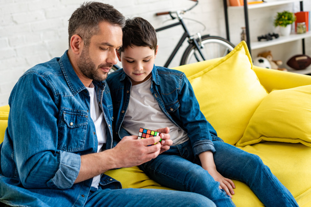 πατέρας και γιός κάθονται στον καναπέ και παίζουν με το παιχνίδι Cube στο σπίτι - Φωτογραφία, εικόνα