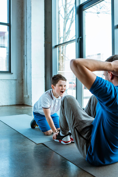 son shouting while helping father doing sit up exercise at gym - Фото, зображення
