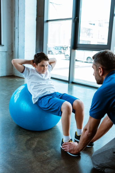 vader helpen zoon zittend op fitness bal en doen zitten oefening op sportschool - Foto, afbeelding