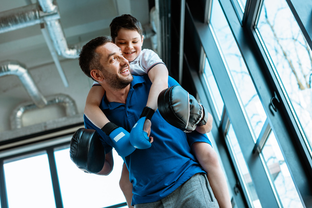 father giving piggyback ride to smiling son in boxing gloves at gym - Fotoğraf, Görsel