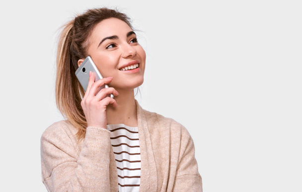 Horizontal indoor shot of young pretty woman smiling and talking on cellphone to her friend, looking up cheerful and happy, posing on white studio background. Real human emotions - Photo, Image