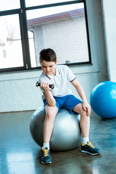 cute boy sitting on fitness ball and working out with dumbbell at gym - Foto, Bild