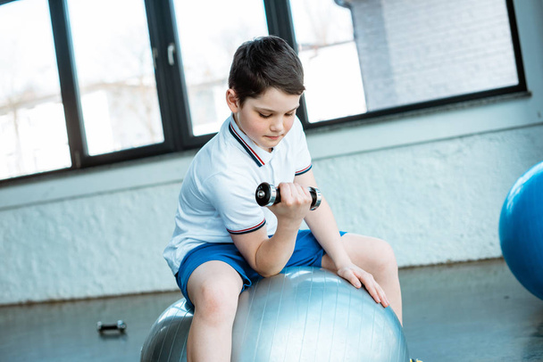 cute boy sitting on fitness ball and working out with dumbbell at gym - Photo, Image