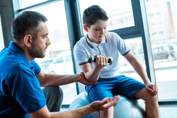 padre guapo ayudando a su hijo a hacer ejercicio con mancuerna en el gimnasio
 - Foto, Imagen