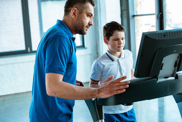 father looking at screen on treadmill while son running at sports center - Photo, Image