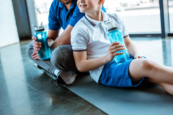 περικομμένη θέα του πατέρα και του γιου που κάθονται στο χαλάκι Fitness με αθλητικά μπουκάλια στο γυμναστήριο - Φωτογραφία, εικόνα