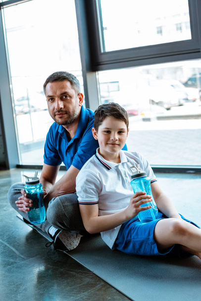 πατέρας και γιός κάθονται στο χαλάκι Fitness με μπουκάλια σπορ και κοιτάζοντας την κάμερα στο γυμναστήριο - Φωτογραφία, εικόνα