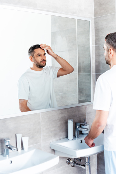 красивый мужчина смотрит на зеркало и регулирует прическу в ванной комнате
 - Фото, изображение