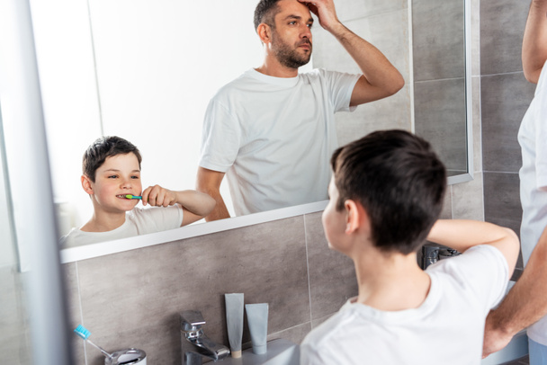 son brushing teeth while father adjusting hairstyle in bathroom - Fotó, kép