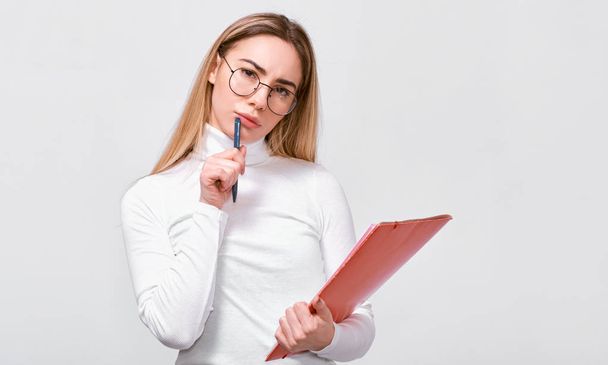 Indoor image of serious young businesswoman wearing white blouse and round trendy eyeglasses with red folder in hand, with blank copy space for your text.  - Φωτογραφία, εικόνα