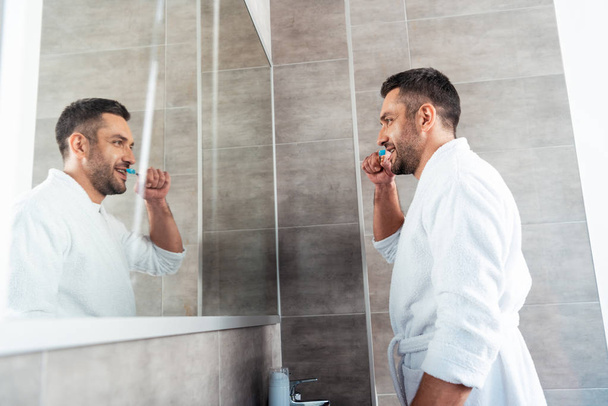 όμορφος άντρας με λευκό μπουρνούζι βουρτσίζει τα δόντια στο μπάνιο κατά τη διάρκεια του πρωινού ρουτίνας - Φωτογραφία, εικόνα