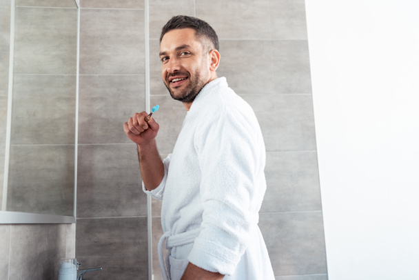 красивый мужчина в халате смотрит в камеру и чистит зубы во время утренней рутины.
 - Фото, изображение