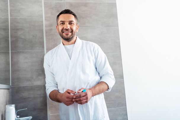 όμορφος χαμογελαστός άντρας με λευκό μπουρνούζι με οδοντόβουρτσα κοιτάζοντας την κάμερα στο μπάνιο - Φωτογραφία, εικόνα