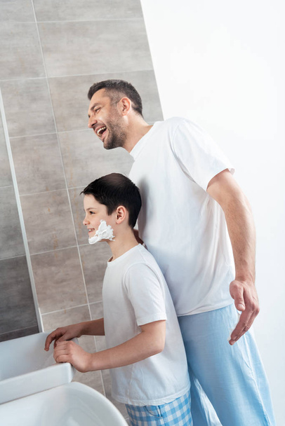 smiling son with shaving cream on face near happy father in bathroom - Фото, изображение
