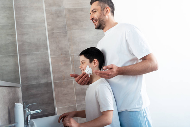 father applying shaving cream on face of son in bathroom - Photo, Image