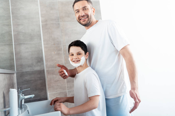 smiling father near son with shaving cream on face in bathroom - Foto, afbeelding