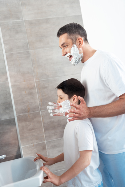 smiling father applying shaving cream on face of son in bathroom in morning - Zdjęcie, obraz