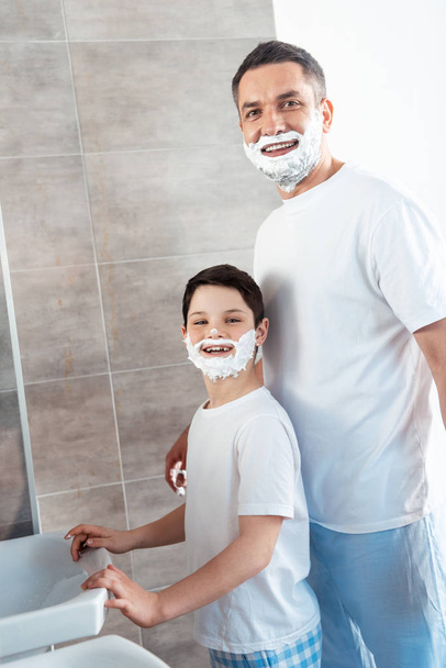 happy son and father with shaving cream on faces looking at camera in bathroom - Fotó, kép