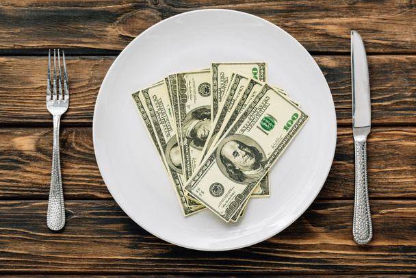 вид сверху на белую тарелку с долларовыми банкнотами рядом с ножом и вилкой на коричневой деревянной поверхности
 - Фото, изображение