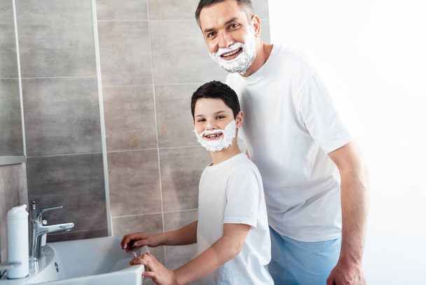 smiling son and father with shaving cream on faces looking at camera in bathroom - Photo, Image