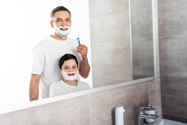 smiling son and father with shaving cream on faces looking at camera in bathroom - Fotografie, Obrázek