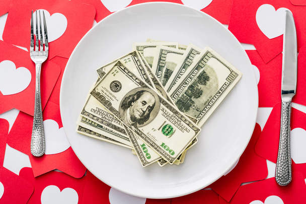 top view of dollar banknotes on white plate near knife and fork on red paper cut cards with hearts symbols - Photo, Image