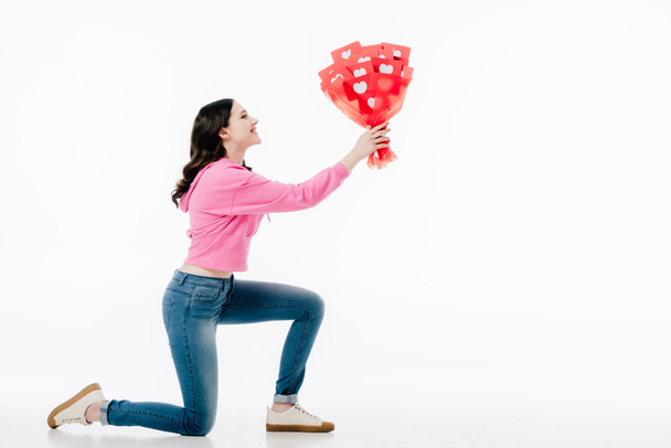 side view of young woman standing on knee and holding bouquet of red paper cut cards with hearts symbols isolated on white - Foto, immagini