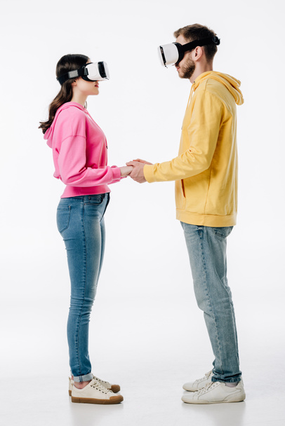 young man in woman in blue jeans and hoodies holding hands while using virtual reality headsets on white background - Foto, immagini