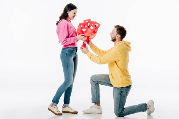 young man standing on knee and gifting bouquet of red paper cut cards with hearts symbols to smiling girl on white background - Foto, afbeelding