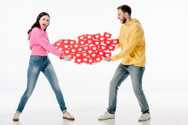 young couple in blue jeans and hoodies quarreling while holding red paper cards with hearts symbols on white background - Φωτογραφία, εικόνα